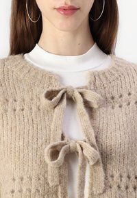 Béžový pletený cardigan s jemnou texturou, který má kulatý výstřih, dvě textilní šňůrky vpředu a složité detaily v prošívání.