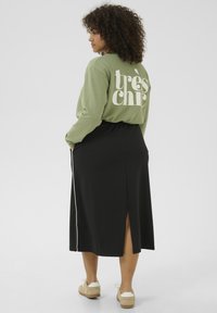 Chemise à manches longues vert olive avec le texte blanc "très chic", associée à une jupe noire avec une fente sur le côté, complétée par des chaussures beiges.