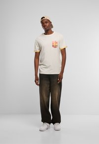 Camiseta beige claro con un bolsillo de patrón colorido, rematado en amarillo en las mangas, combinada con pantalones de pierna ancha oscuros y ligeramente desgastados, y zapatillas blancas.