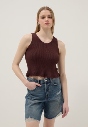 Frau, die ein ärmelloses, geripptes, dunkelbraunes Crop-Top und hoch taillierte, ausgefranste Denim-Shorts trägt, steht vor einem einfarbigen Hintergrund.