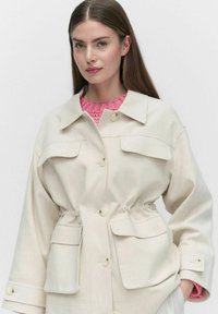 Krämfärgad jacka med dragsko i midjan, stora framfickor och krage. Har en slät yta och knappstängning.