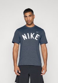 Tumši zils Nike T-krekls ar īsām piedurknēm, kas rotāts ar lielu baltu "NIKE" logotipu uz krūtīm un mazu baltu Nike swoosh uz pleca.