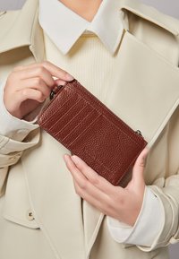 Braune Lederbörse mit strukturierter Oberfläche, ausgestattet mit Kartenfächern, Reißverschluss und einer schlanken rechteckigen Form. In der Hand gehalten gegen einen hellen Mantel.