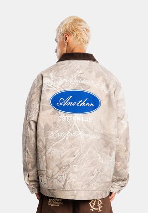 Person med kort blondt hår iført beige camouflagejakke med blåt ovalt logo og hvid tekst på ryggen, stående mod en ensfarvet lys baggrund.