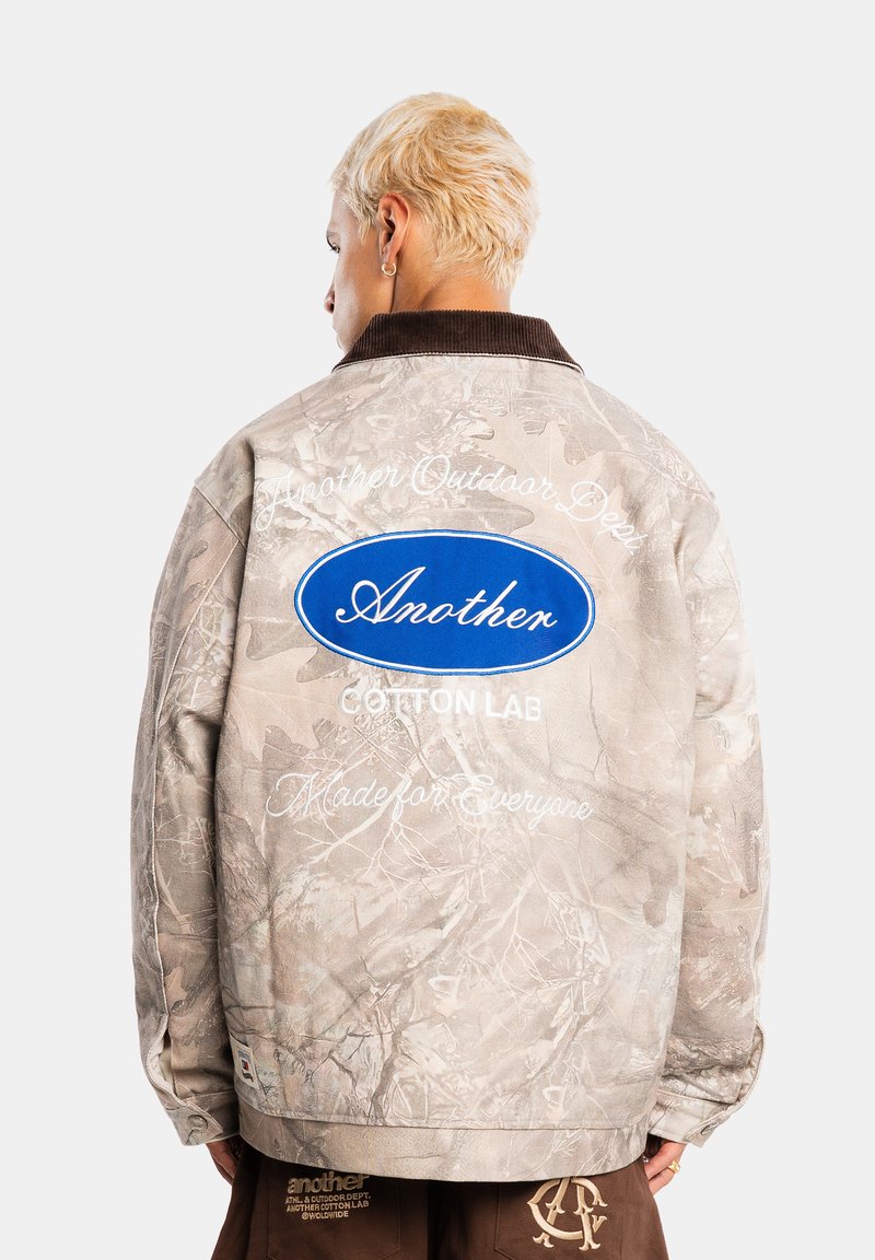 Personne aux cheveux blonds courts portant une veste camouflage beige avec un logo ovale bleu et un texte blanc dans le dos, debout devant un fond clair uni.