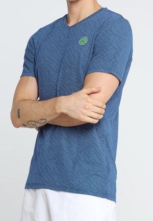 Man die een blauw overhemd met korte mouwen en patroon draagt, met een groen logo op de borst, witte short, armen gekruist, toont tatoeages op de linker onderarm.