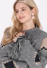 Sivi pleteni pulover s uzorkom u bež i mornarsko plavoj boji, s detaljima od resica. Model nosi okrugle naušnice i prstenje.