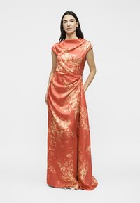 Γυναίκα που φορά φορεμα από κοραλλί σατέν μήκους μέχρι το πάτωμα με χρυσά λουλουδένια σχέδια και ντραπέ ντεκολτέ, που στέκεται μπροστά σε απλό φόντο.