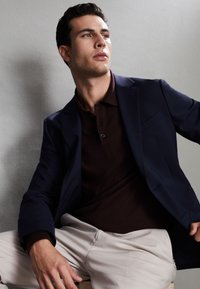 Jonge man zittend, draagt een navy blazer, donkerbruin poloshirt en lichtbeige broek, kijkt iets naar rechts tegen een effen muur.