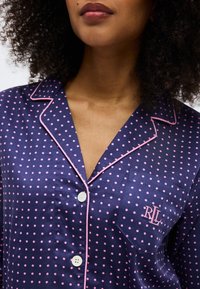 Γυναίκα που φοράει σατέν πιτζάμες σε σκούρο μπλε με ροζ πουά, ροζ περιθώρια, λευκά κουμπιά και το κέντημα "RLL" στην τσέπη του στήθους.