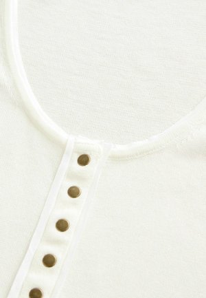 Weißes Baumwolloberteil mit Rundhalsausschnitt und einer Knopfleiste mit fünf messingfarbenen Knöpfen. Der Stoff hat eine feine Textur.