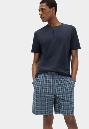 Man wearing a navy short-sleeve henley shirt and blue plaid drawstring shorts, standing with hands in pockets, looking left.