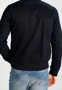 Man wearing a fitted black jacket and light blue jeans, shown from the back with arms relaxed at sides.