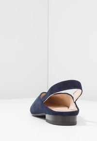 Sko i mørkeblå ruskind med lav sort hæl og åben bagpå, med en glat inderside og et buet remdesign.