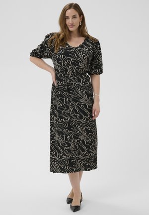 Moteris su ilgais rudais plaukais, vilkinti juodą midi suknelę su smėlio spalvos abstrakčiais raštais, pusiau rankovėmis ir juodais smailiais batais.