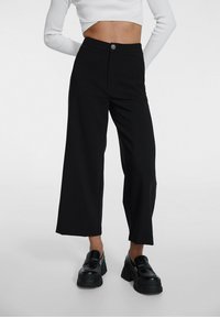 Zwarte wijde broek met een hoge taille, voorzien van een knoopsluiting aan de voorkant, een gladde textuur en een minimalistisch design. Gepaard met chunky zwarte schoenen.