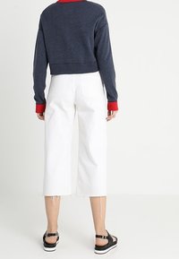 Zkrácený námořnický svetr s červeným výstřihem, spárovaný s bílými kalhotami s širokými nohavicemi. Černé sandály mají nastavitelné pásky a bílé podešve.