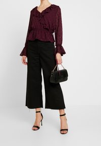 Blusa bordeaux con volant e motivo sottile; pantaloni neri a gamba larga; décolleté neri con cinturini; borsa nera a tracolla con catena.