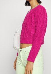 Knallpinkes Kabelstrickpullover mit kürzerem Schnitt und langen Ärmeln, kombiniert mit hellgrünen, hoch taillierten Jeans und einer kleinen weißen Tasche.
