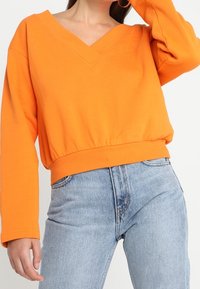 Sweatshirt orange surdimensionné avec un décolleté en V profond, ourlet côtelé et manches longues, associé à un jean en denim bleu clair.