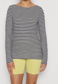 Osoba ubrana w czarną i białą poziomo paskowaną koszulę z długim rękawem oraz żółte dopasowane szorty do kolan, stojąca na jednolitym tle.