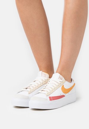 Pieds portant des baskets basses blanches avec des détails Nike swoosh orange et rouge sur un fond blanc uni.