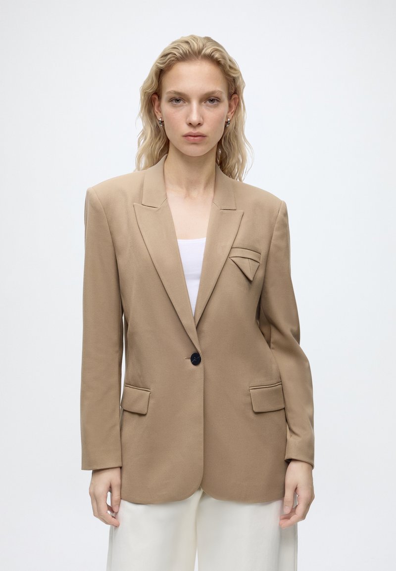 Kvinde med langt blond hår iført beige skræddersyet blazer over hvid top og hvide bukser, stående mod en ensfarvet lys baggrund.
