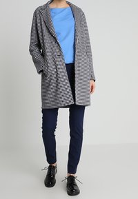 Schwarz-weiß karierter Mantel mit lockerer Passform, kombiniert mit einem blauen Oberteil und marineblauen Hosen. Glänzende schwarze Schuhe runden das Outfit ab.