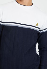 Granatowy sweter o teksturowanym splocie z białymi i szarymi paskami, ribbowanym okrągłym dekoltem oraz małym żółtym logo ptaszka na piersiach.