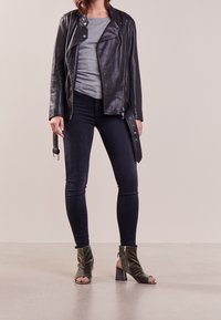 Kobieta w czarnej skórzanej kurtce, szarej bluzce, czarnych dopasowanych dżinsach i zielonych, otwartych na palce, obcasach do kostek stoi na polerowanej podłodze.