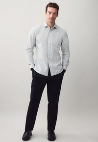 Camicia a maniche lunghe con bottoni in azzurro chiaro con sottili righe verticali, abbinata a pantaloni scuri e scarpe nere. Presenta una tasca sul petto.