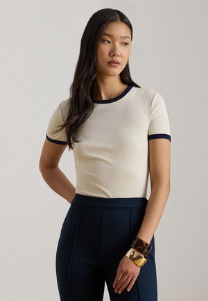 Tricou crem cu mâneci scurte și tiv bleumarin, design ajustat, textură moale. Asortat cu pantaloni închiși la culoare, croiți, și brățări suprapuse, inclusiv cu model de carapace de țestoasă.