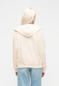 Γυναίκα με μακριά ξανθά μαλλιά, που φοράει ανοιχτό μπεζ φούτερ με κουκούλα και ανοιχτό μπλε τζιν, γυρισμένη με την πλάτη σε απλό λευκό φόντο.