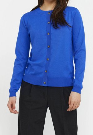 Femme portant un cardigan boutonné bleu vif et un pantalon noir, montrée des épaules aux hanches sur un fond uni.