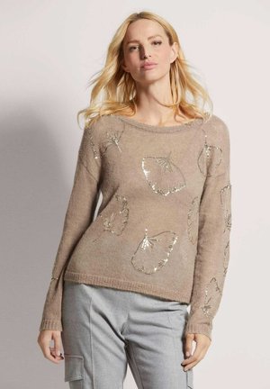 Beige trui met lange mouwen, met pailletten vlinderpatronen. Zachte textuur, ronde halslijn en ribbelrand. Gecombineerd met lichtgrijze broeken.