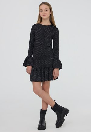 Ragazza con lunghi capelli biondi che indossa un vestito nero con pois, maniche svasate e orlo arricciato, abbinato a stivaletti neri.
