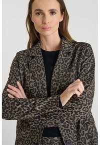 Blazer en imprimé léopard en tissu marron et noir. Revers crantés, deux boutons et manches longues avec poignets repliés. Détails de motif texturé.