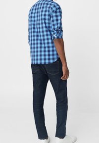 Niebieska i czarna koszula w kratę z podwiniętymi rękawami, zestawiona z ciemnoniebieskimi dżinsami i białymi trampkami, widok z tyłu.