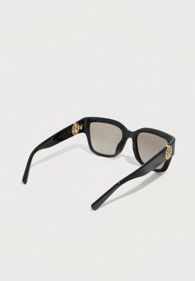 Γυαλιά ηλίου με μαύρο τετράγωνο πλαίσιο, καφέ φακούς με σκίαση και χρυσά κυκλικά διακοσμητικά λογότυπα στα πλαινά, εμφανιζόμενα σε λευκό φόντο.