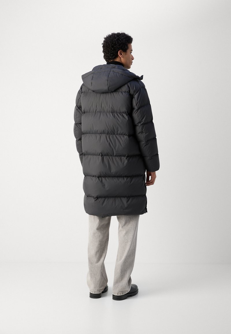 Abrigo de plumas negro con capucha, con un diseño acolchado, de larga longitud y con aberturas laterales para facilitar el movimiento, combinado con pantalones gris claro.