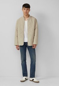 Beige gewatteerde jas met een staande kraag, gedragen over een witte v-hals tshirt, gecombineerd met blauwe spijkerbroeken en witte sneakers met zwarte accenten.
