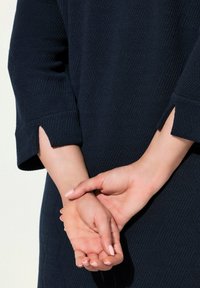 Tyrkysové šaty s texturovaným vzorem a tříčtvrtečními rukávy s rozparky. Ruce jsou sepnuty před tělem.