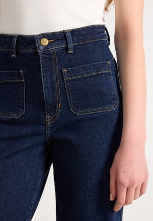 Högmidjade mörk denimjeans med gula sömmar, med framfickor och guldknappstängning, slät textur och rak passform.