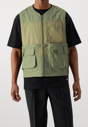 Homme portant un gilet utilitaire vert bicolore avec plusieurs poches avant par-dessus un t-shirt noir à manches courtes et un pantalon noir.