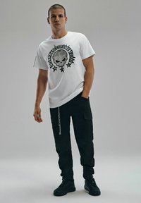 Biała koszulka z krótkim rękawem z czarnym nadrukiem czaszki i gwiazdy. W zestawie z czarnymi spodniami cargo i czarnymi trampkami. Prosty design i fason.