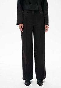 Zwarte wijde broek met een op maat gemaakte pasvorm en een soepele textuur, voorzien van voorplooien en een hoge taille. Gecombineerd met puntige zwarte schoenen.