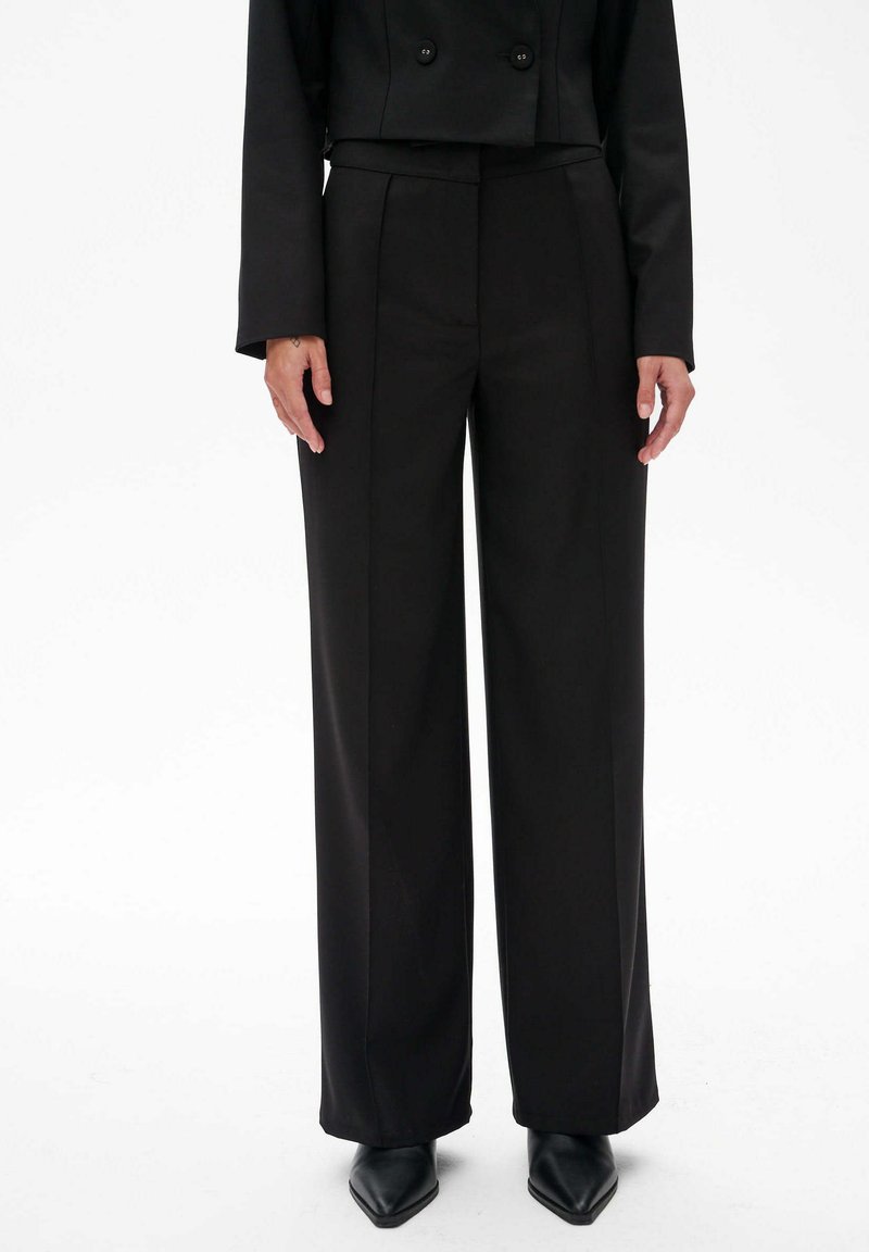 Zwarte wijde broek met een op maat gemaakte pasvorm en een soepele textuur, voorzien van voorplooien en een hoge taille. Gecombineerd met puntige zwarte schoenen.