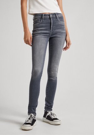Slim-fit vervaagde grijze jeans gedragen met zwart-witte sneakers en een wit mouwloos topje, staand tegen een effen achtergrond.
