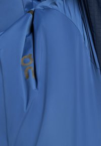 Veste bleue avec une poche latérale zippée et un petit logo noir sur la manche près de la poche.