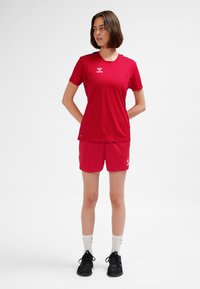 Rode sportieve shirt- en shortset gemaakt van lichtgewicht stof, met een V-hals, logo en contrasterende schouderaccenten. Draagt zwarte schoenen.